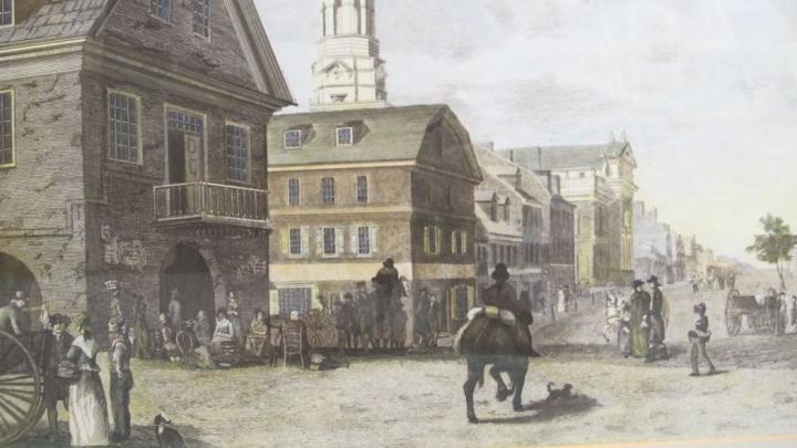 Historical town street with pedestrians, horse riders, carts, and buildings surrounding a public square.