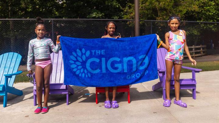 Children at a community pool display a towel with The Cigna Group logo, representing support for youth and community well-being.