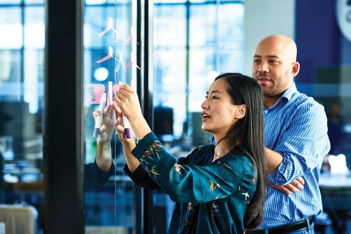 Two employees collaborate in an office, placing notes on a glass wall during a planning or brainstorming session.