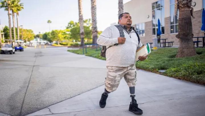 Person wearing a prosthetic leg walks along a sidewalk carrying documents near residential buildings.