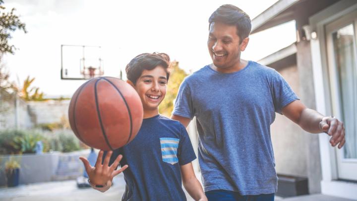 Young person holds a basketball while walking outdoors beside an adult in a residential neighborhood.