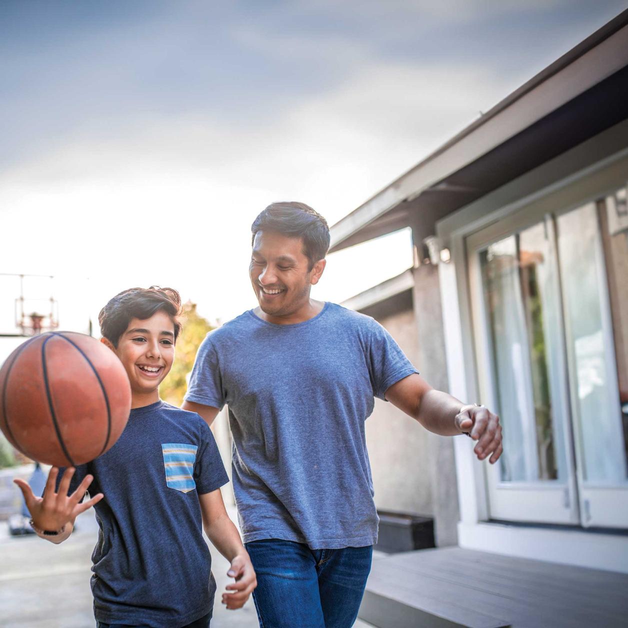 Adult and child outdoors beside a home as the child balances a basketball on one hand.