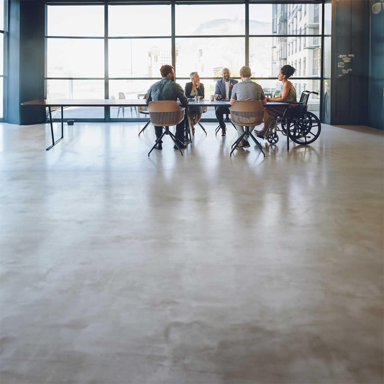 Employees participate in a workplace meeting in an accessible conference room, including a wheelchair user.  More descriptive: