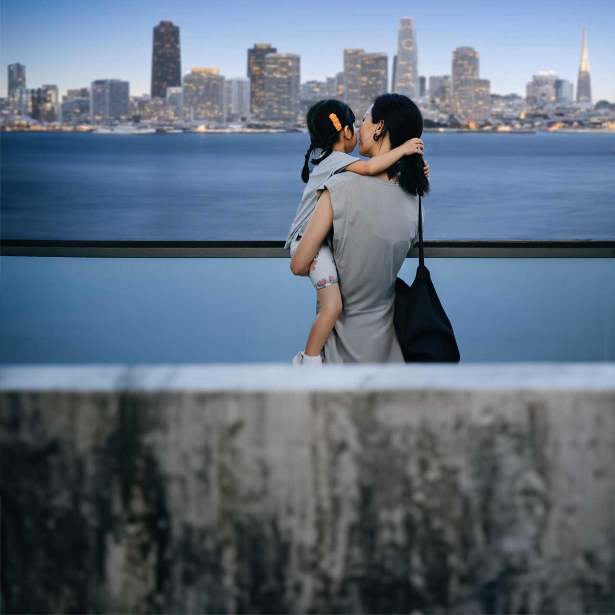 Rear view of a person carrying a child beside a waterfront, overlooking a city skyline across the water at dusk.