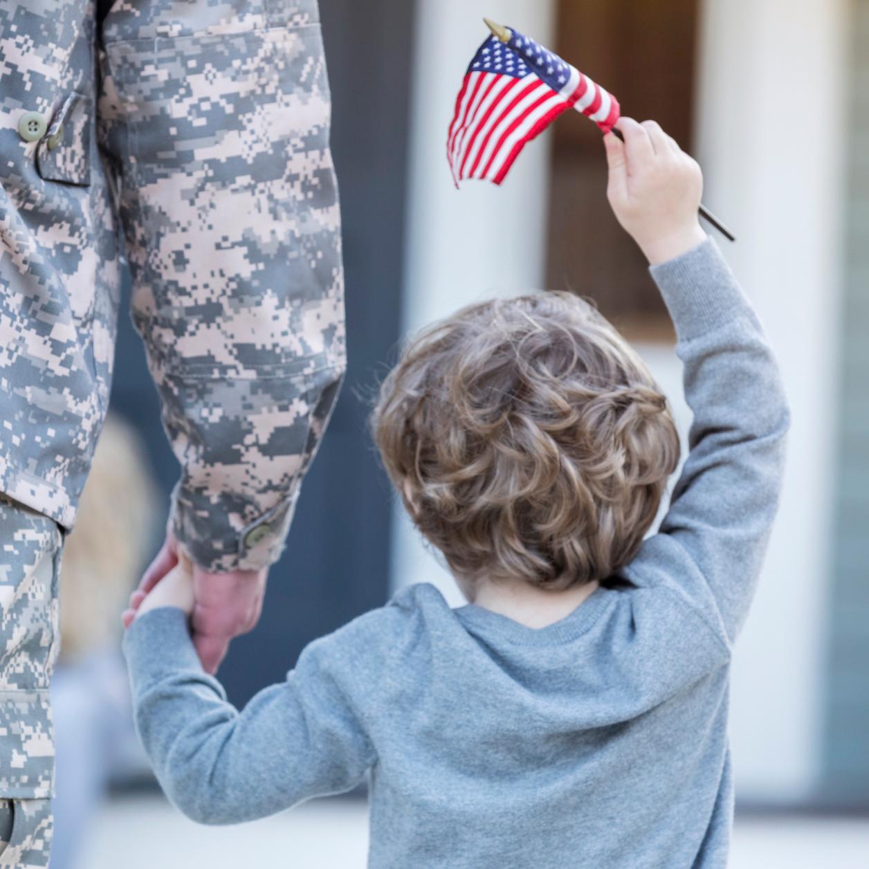 Child holds a small American flag while walking hand in hand with a person wearing a military uniform.
