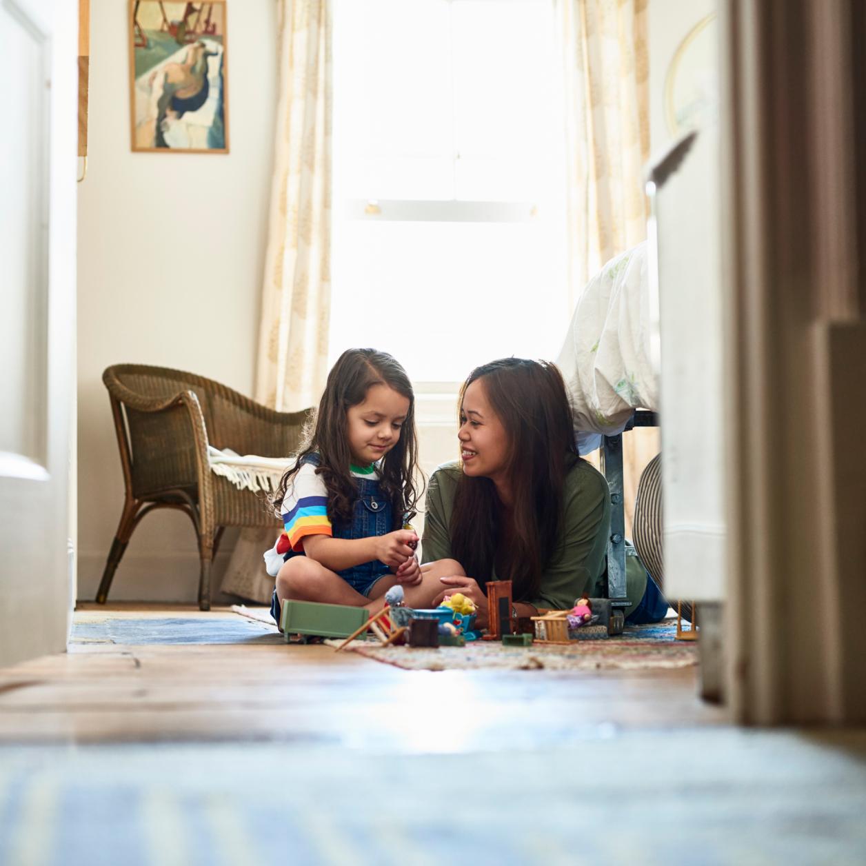 Adult and child sitting on the floor of a home, playing together with toys in a bright living space.