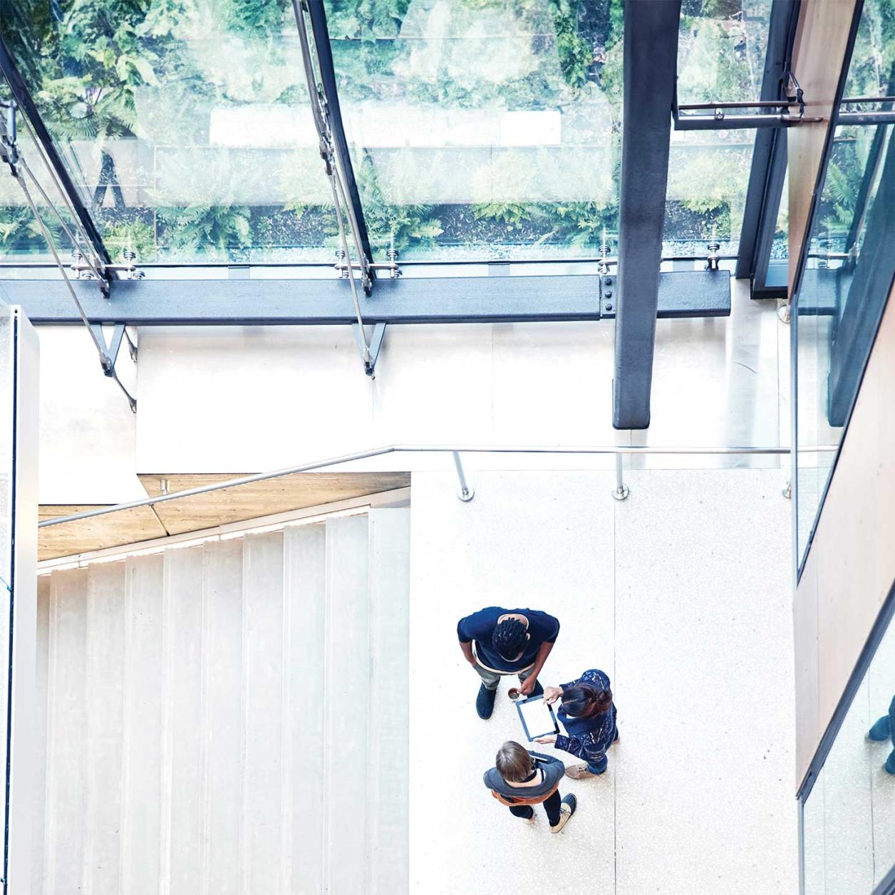Overhead view of three people standing in a bright office atrium, reviewing information on a tablet near a staircase and large glass windows.