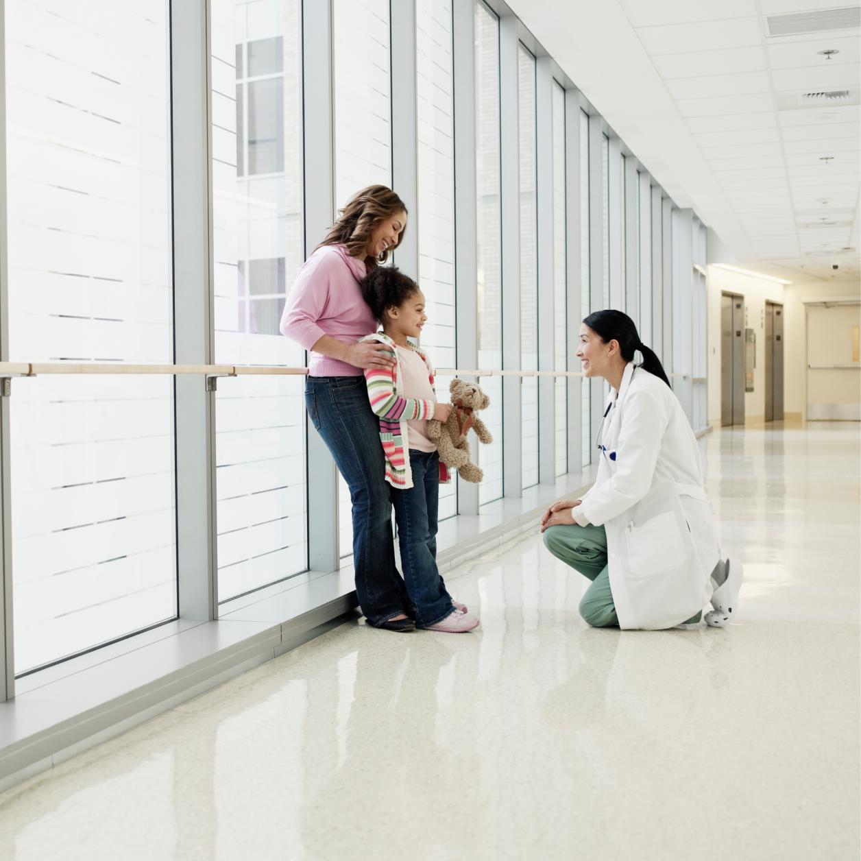 Healthcare professional kneels to speak with a child holding a stuffed toy while another adult stands nearby in a hospital corridor.